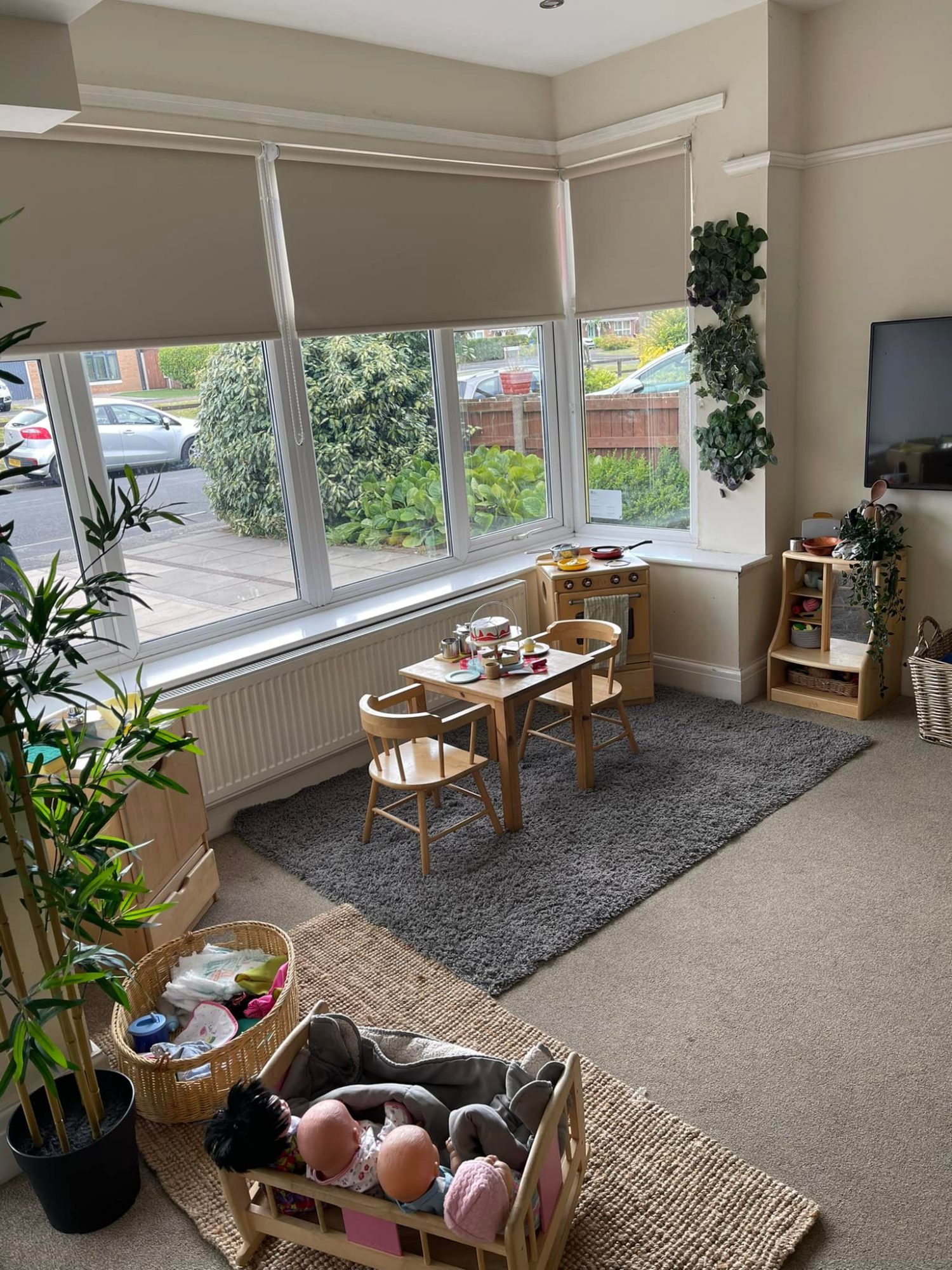 Small wooden children's table and chairs arranged with toy dishes on a gray rug by a bay window in a living room; toy stove, basket of dolls and potted plants nearby.