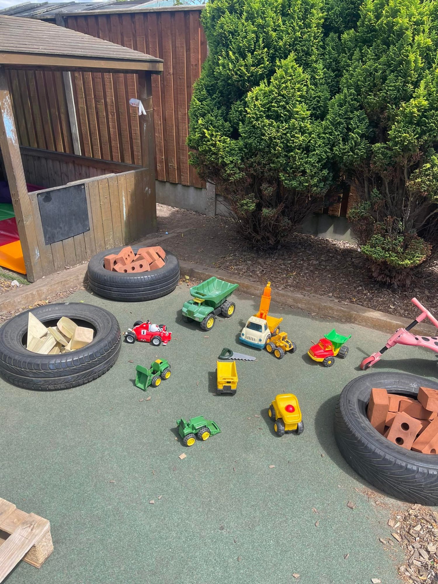 Several small toy trucks and vehicles are scattered across a rubber play surface beside three tires filled with wooden blocks and bricks, near a wooden play hut and shrubs.