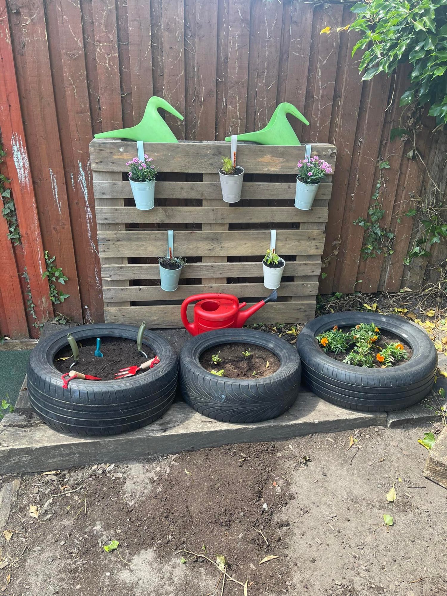 Wooden pallet holds six small hanging potted flowers; three tires serve as planters (one with marigolds); a red watering can rests before a brown fence in a backyard.