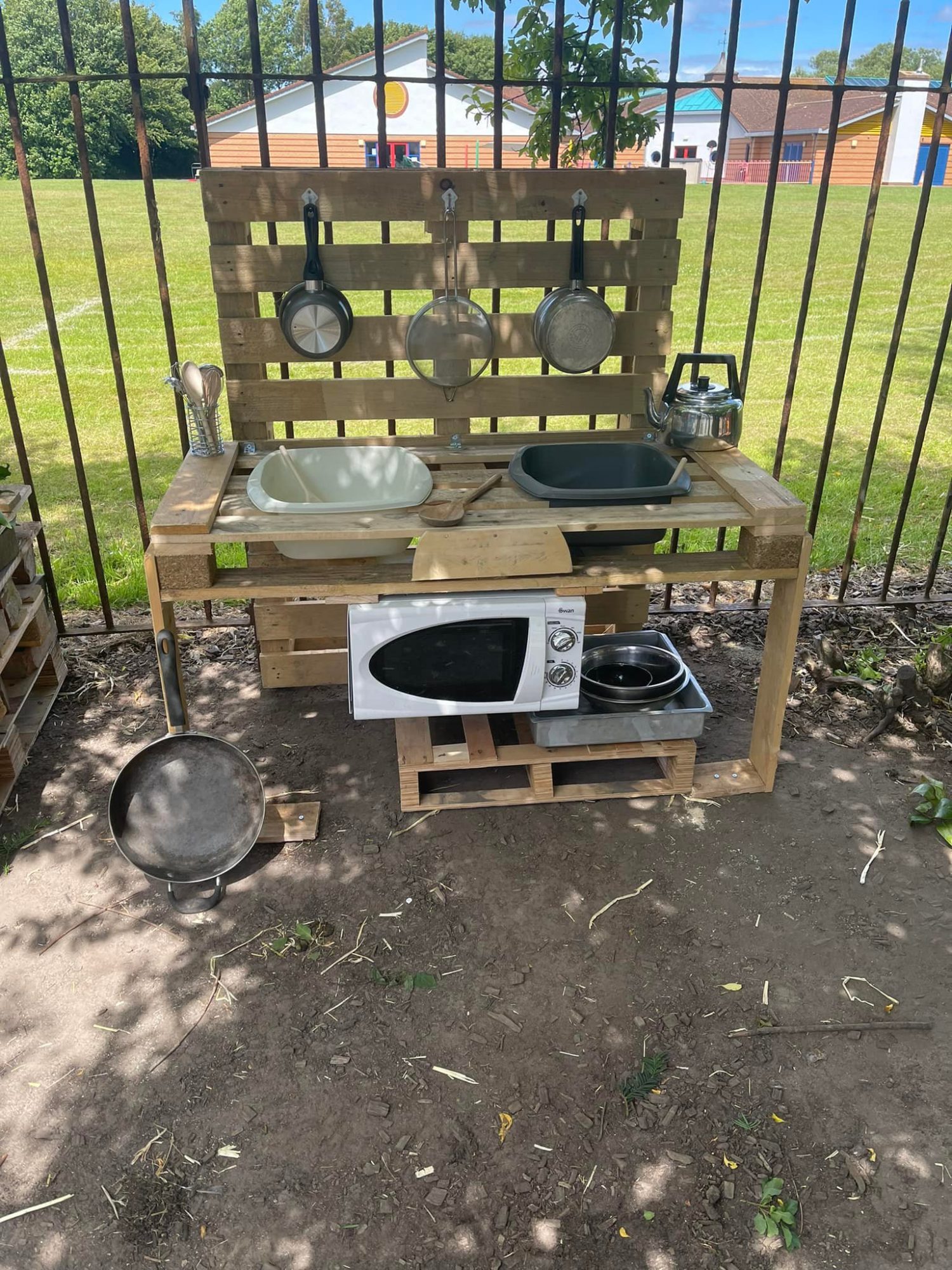 Wooden pallet play-kitchen arranged with two sinks, hanging pans and utensils; microwave and kettle on shelves; positioned outdoors against a metal fence with grassy field and buildings beyond.