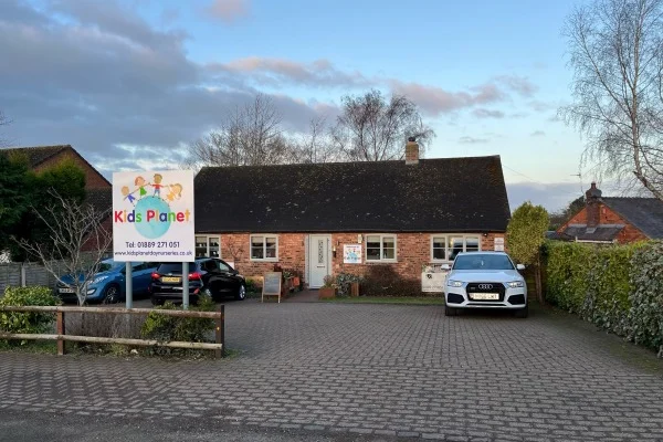 Single-storey brick building with a "Kids Planet" sign; cars parked in the paved driveway, low hedges and bare trees around, under a clear late-afternoon sky.