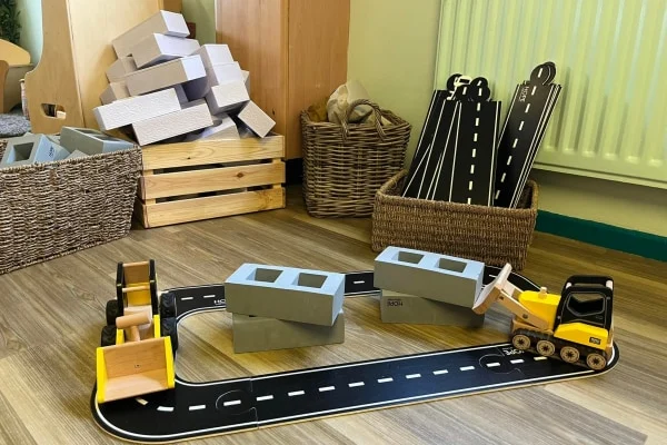A toy road track loops around wooden construction vehicles pushing gray block pieces, set on a classroom wood floor amid wicker baskets, a wooden crate and stacked foam blocks. No legible text.