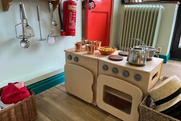 Wooden play stove holds a kettle, pots and copper cups on faux burners; metal utensils hang on the wall, wicker baskets sit nearby, radiator and red door in the background. Text on extinguisher: "Foam"