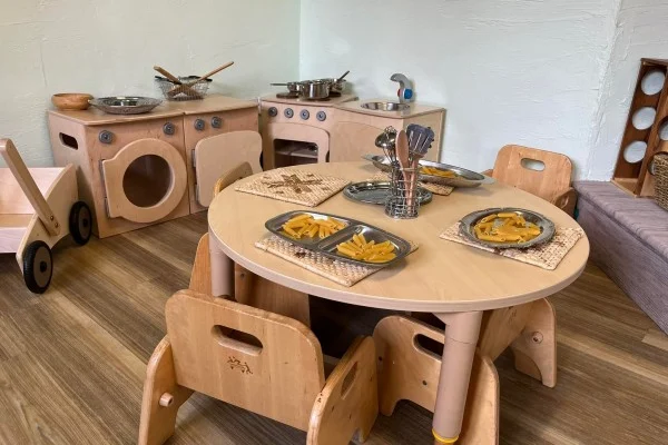 Round wooden children's table holds metal plates of pretend pasta and utensils; child-sized chairs tucked under; a wooden play kitchen with stove, sink, and toy pots sits against the wall.