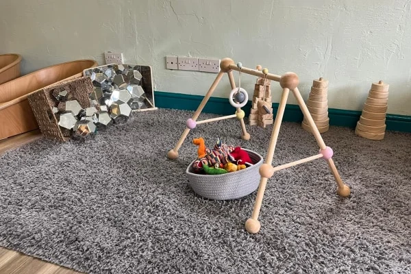 Wooden baby gym stands over a basket of plush toys on a gray rug, in a corner playroom with a mirrored sensory board, stacked wooden cones, wall and teal skirting.