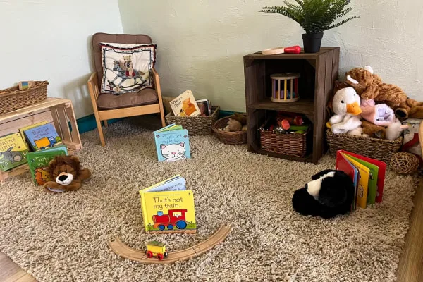 Yellow board book sits on a shag rug beside a curved wooden toy train track; stuffed animals, baskets, a small chair and wooden shelf with more books surround it.