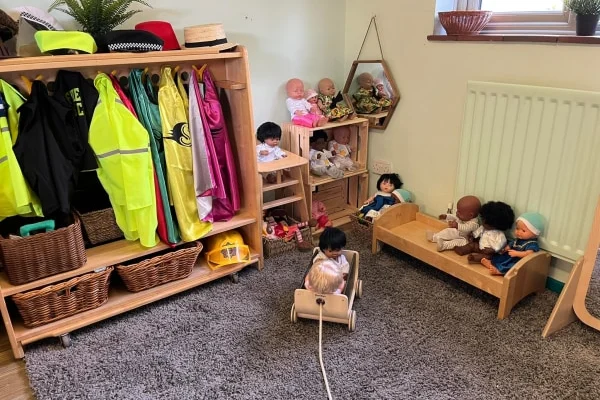 A small doll sits in a wooden pull-cart being pulled across gray carpet toward a low wooden bed holding several dolls; shelving nearby displays dress-up coats and woven baskets.