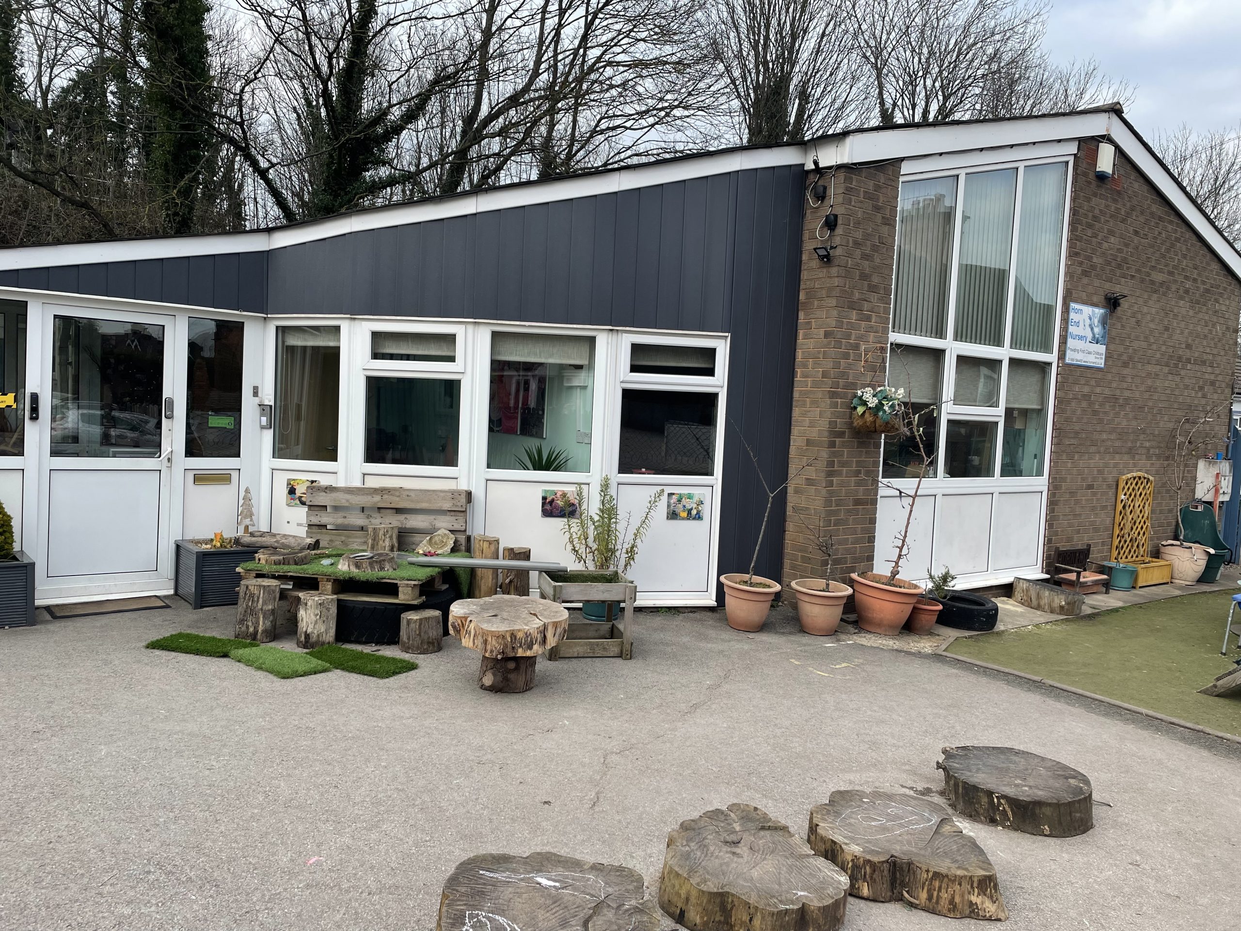 Single-story nursery building displays wooden log stools and a pallet bench outside; potted plants and a hanging basket flank the entrance; paved courtyard and bare trees form the surrounding setting.