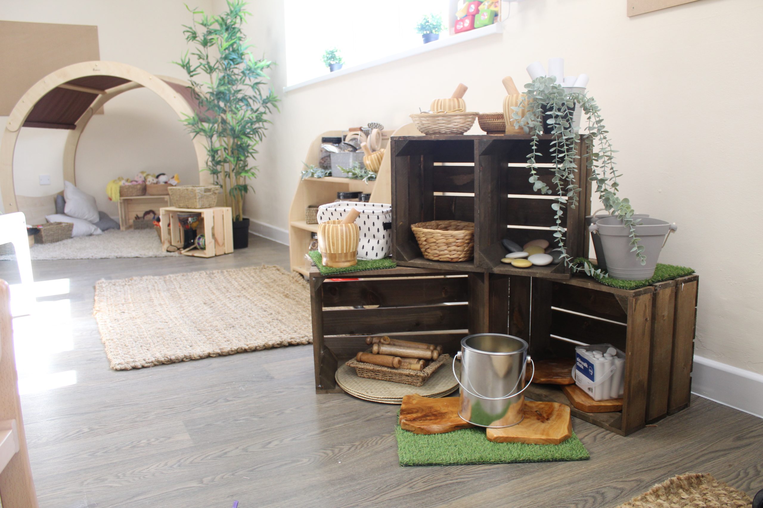 Stacked wooden crates hold baskets, toy kitchen tools, a metal pail and trailing plant, forming a play-storage display in a bright children's room with rug and arched cozy nook.