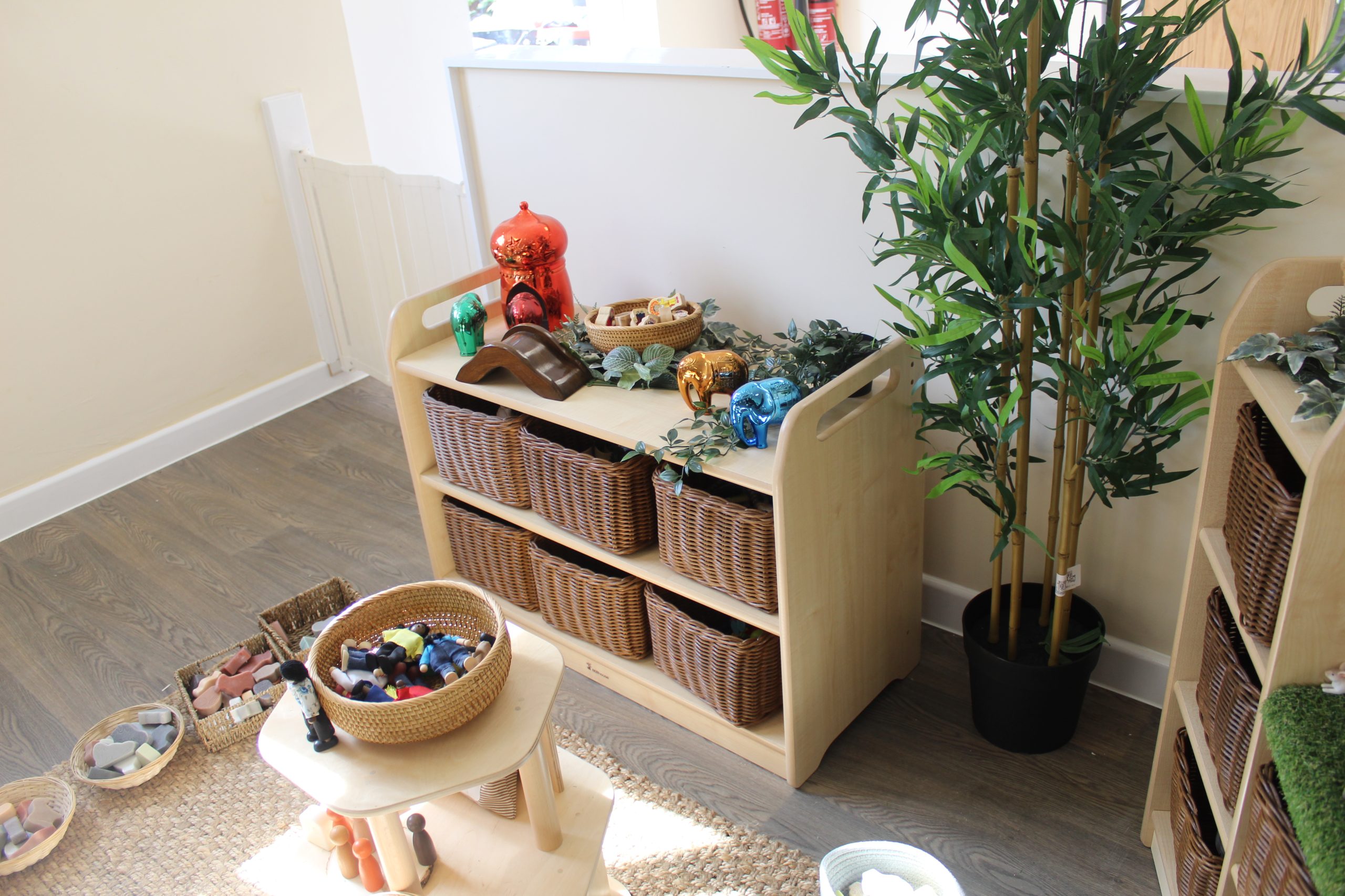 Wooden low shelving unit holds six wicker baskets and assorted toys and decorative elephant figurines atop; placed against a pale wall beside a tall potted bamboo plant in a playroom.