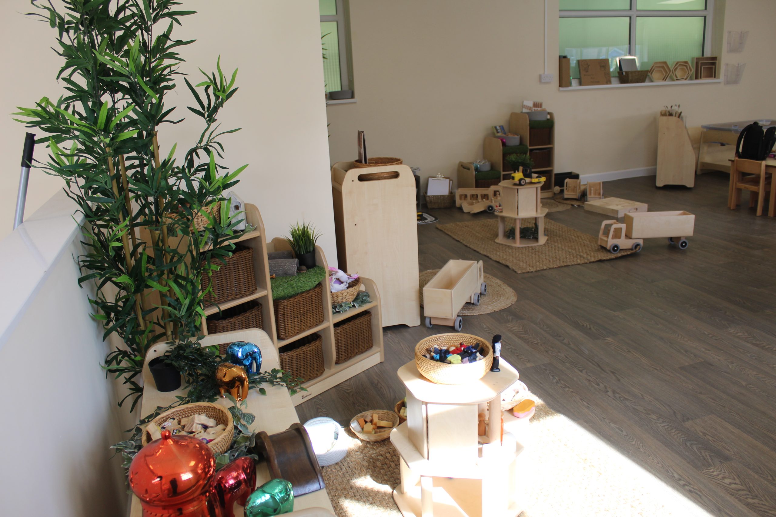 Wooden toys and low shelving are neatly arranged on rugs and a hardwood floor, bathed in sunlight inside a tidy preschool playroom with wicker baskets and a tall potted plant.