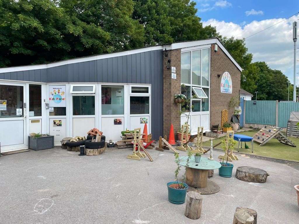 Single-story children's center building framed by trees, with outdoor play setup — wooden stumps, spool tables, ramps, cones and plant pots — arranged on a paved courtyard under blue sky.