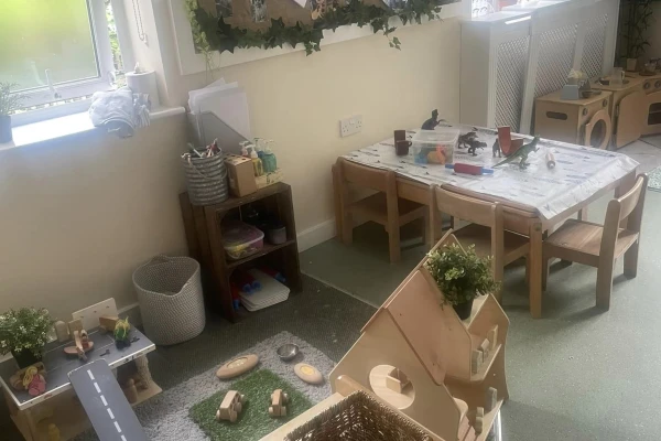 Wooden dollhouse sits beside a small table with child-sized chairs; toy cars on a rug road, craft supplies on a shelf, and a play kitchen against the sunlit classroom wall.