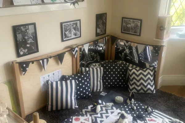 Black-and-white cushions cluster around a patterned playmat; small toys sit on it; triangular bunting and framed black-and-white photos hang in a mirrored corner of a playroom.