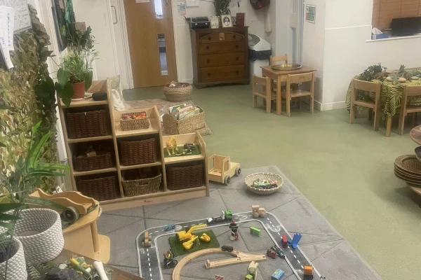 Wooden toy train and car set sprawls across a gray play mat with tracks and vehicles; low shelves, wicker baskets, small tables and plants frame a quiet preschool classroom.