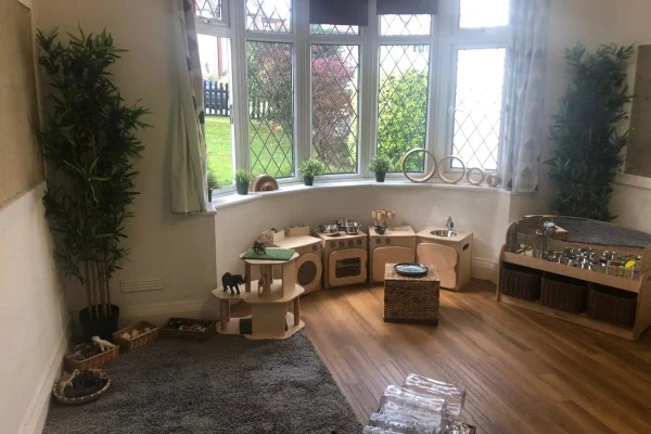 Child-scale wooden play kitchen and shelving arranged along a bay window, stocked with toy dishes and baskets for pretend cooking, situated on hardwood floor with a gray rug and potted plants.