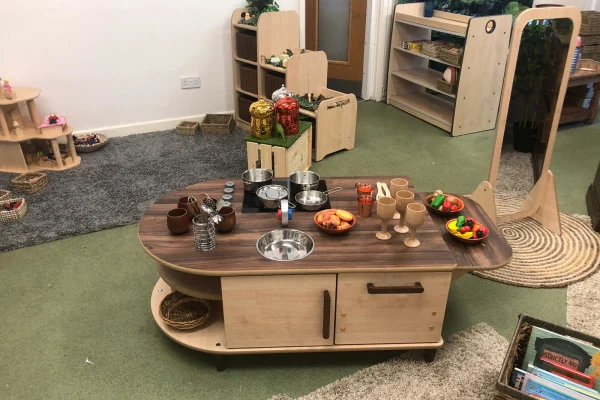 Low wooden play-kitchen island displaying toy pots, pans, bowls, wooden goblets, plastic fruit and jars, arranged for pretend cooking in a carpeted preschool corner with shelves, mirror, and rugs.