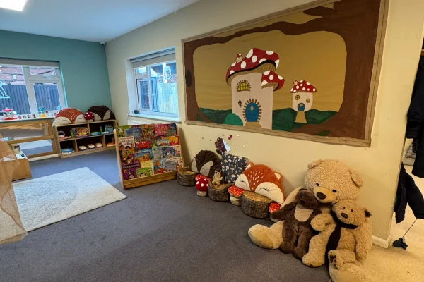 A row of plush toys (bears, foxes) sit against a wall in a carpeted reading corner with low shelves of books and toys beneath windows; a mushroom-house mural above.