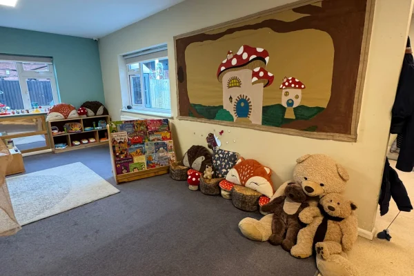 A cluster of stuffed teddy bears sits against a wall beneath a painted mushroom mural, beside low shelves of children's books in a carpeted daycare playroom.