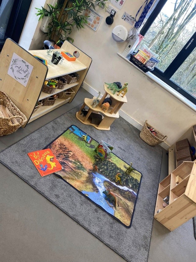 Small play rug holds several toy dinosaurs and a children's dinosaur book, placed on a carpeted floor in a bright classroom corner with low wooden shelves, wicker baskets and a window.