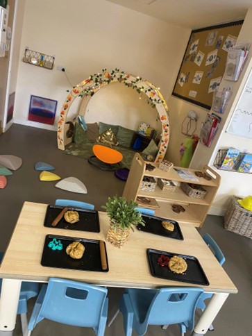 Child-sized table laid with four black trays holding playdough and tools; small potted plant centerpiece; blue chairs; decorated arch shelters a cushioned reading nook in a preschool classroom.
