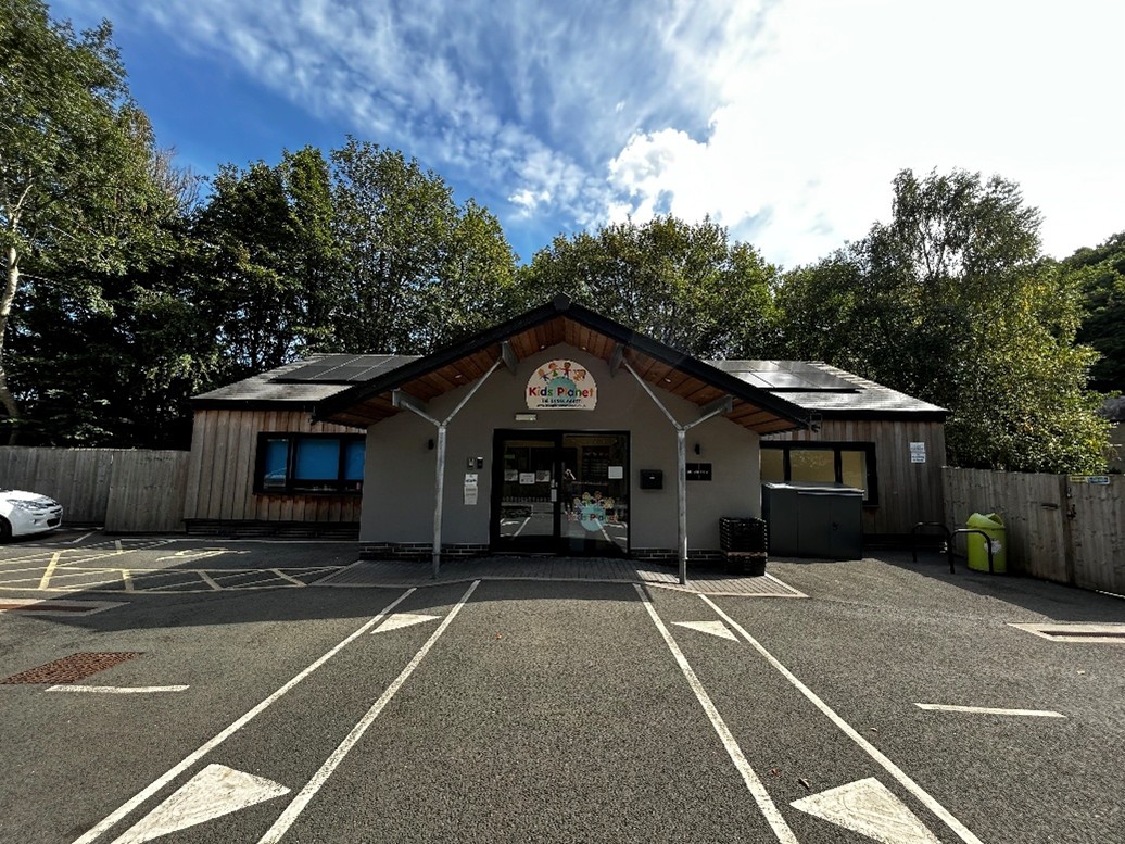 Single-story childcare building with a "Kids Planet" sign, glass entrance door, and covered porch; parking lines and directional arrows lead to the forecourt, surrounded by trees under a blue sky.