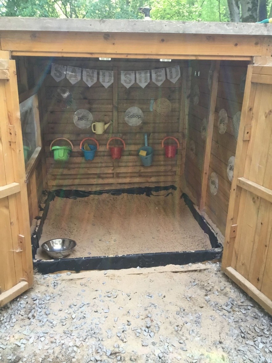 Wooden sand shed contains a shallow sandbox with sand, hanging colorful buckets and play tools, and open double doors revealing a sheltered play area in a leafy backyard.