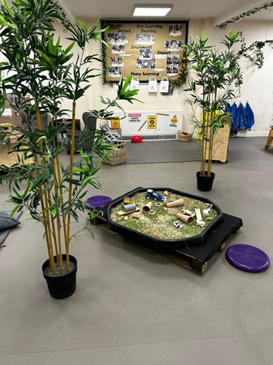 A low sensory play tray filled with shredded material and scattered tubes and toys; flanked by potted bamboo in a classroom with a bulletin board and storage cubbies.