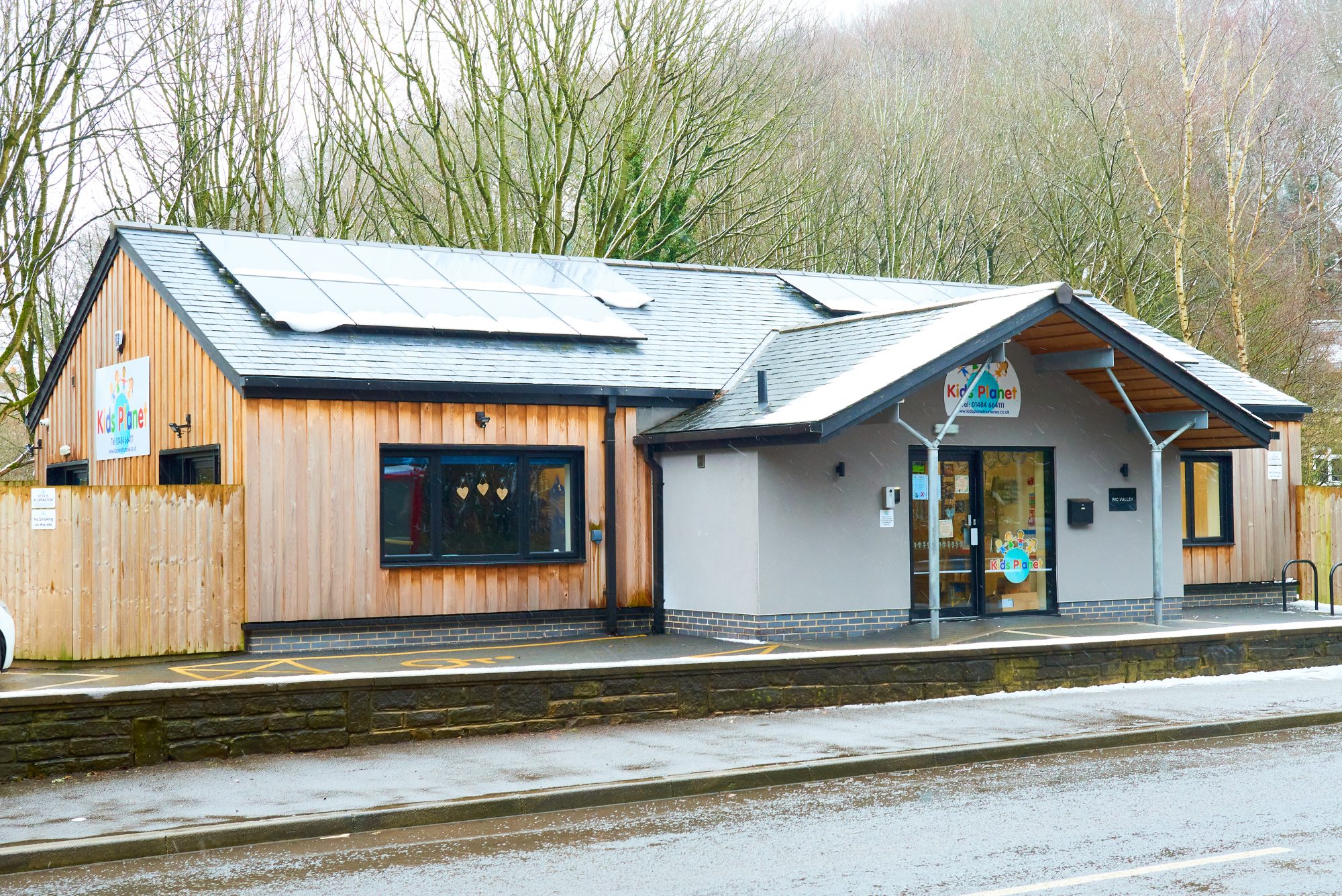 Children's daycare building sits with a light dusting of snow on its roof and solar panels, backed by leafless trees and bordered by a wet road and low stone wall.