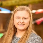 Young woman smiles directly at the camera, long straight blonde hair and gray patterned sweater; colorful triangular bunting and warm wooden background suggest an outdoor festive setting.