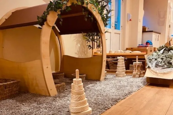 Wooden arch play structures form small shelters on a gray rug, surrounded by wooden stacking cones, baskets and low furniture; a bright, plant-adorned playroom with wooden floor in the background.