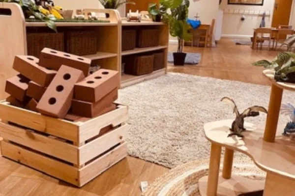 Wooden crate holding brown toy bricks, some stacked and leaning, spilling out; placed on hardwood floor beside rugs, low shelves, plants and small children's tables in a classroom.