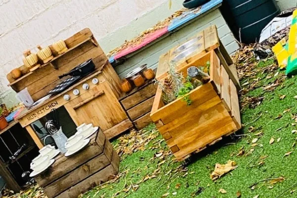 Wooden mud kitchen labeled "MUD KITCHEN" holds toy pots, stacked plates and utensils; adjacent wooden planter contains small herbs; set on artificial grass with fallen leaves against a pale wall.