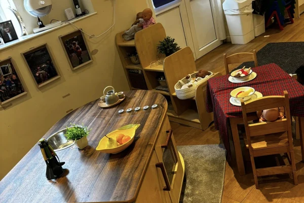 Wooden play kitchen island displays fruit bowl, toy sink, potted plant and toy bottle; nearby table with red-checked cloth holds toy plates and chairs amid dolls and framed photos.