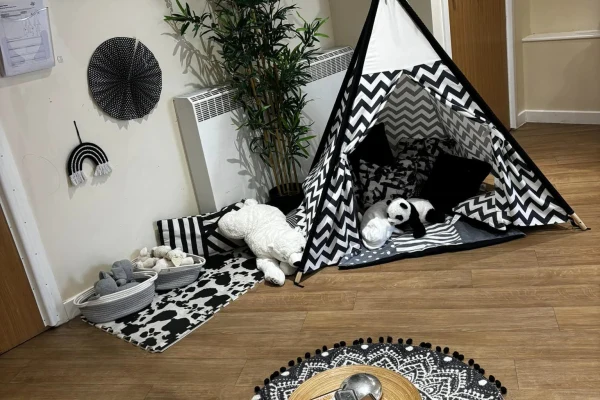Small black-and-white chevron teepee tent houses stuffed panda and lamb, set on a cow-print mat beside baskets of toys; potted plant and circular wall decorations adorn a wooden-floored corner.