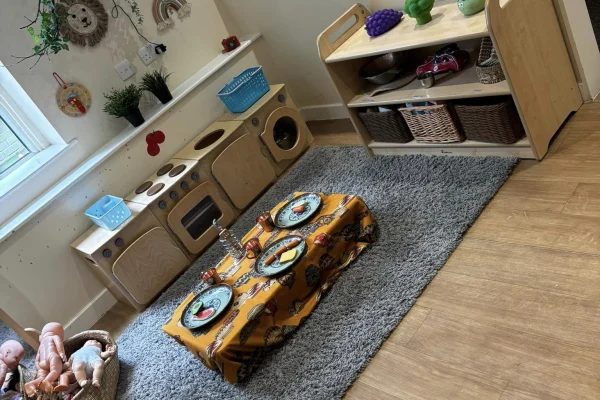 Wooden toy kitchen sits against a wall; a low table draped in yellow cloth holds toy plates and utensils on a gray rug, surrounded by shelves, wicker baskets and dolls.