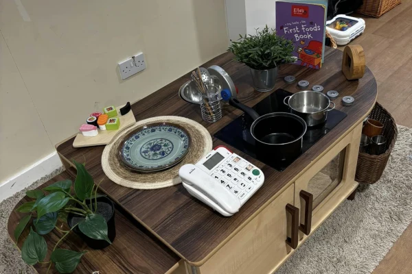 Wooden children's play-kitchen countertop displays toy pots and pans, a decorative plate, a toy phone, utensils, a potted plant and a book on a carpeted floor beside a wall outlet.