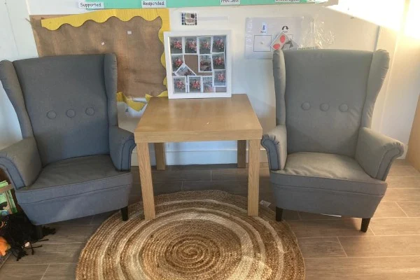 Two gray wingback armchairs flank a small wooden table holding a white photo collage, set on a circular woven rug before a bulletin board on a tiled floor.