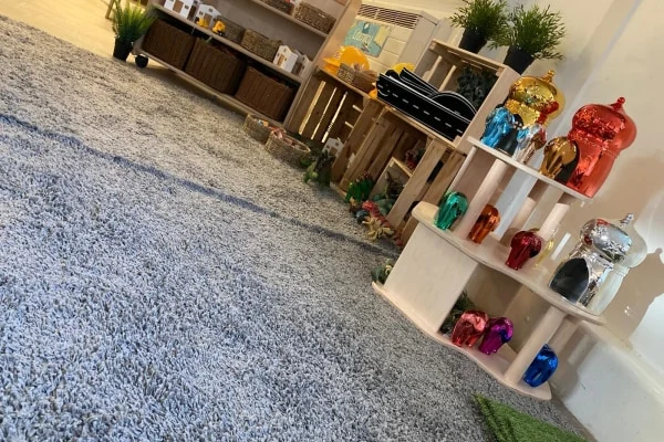 Tiered white display holds assorted shiny metallic pig figurines grouped by color; wooden crates and shelves behind contain baskets and plants; thick gray carpet covers floor in a playroom setting.