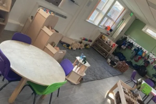 Round activity table with colorful chairs sits amid play stations; wooden shelves hold toys and stuffed animals, a rug defines a play area near windows in a bright preschool classroom.