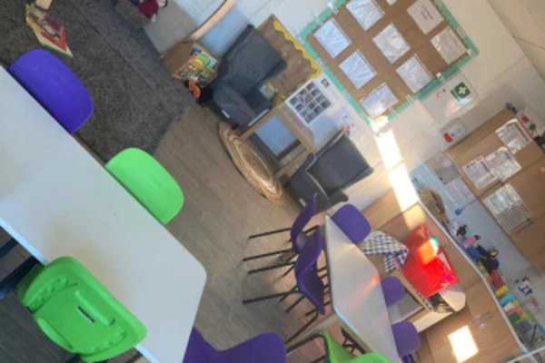 Rows of small green and purple chairs surround white tables, arranged neatly in an empty elementary classroom with a carpeted reading corner, bulletin board, toy bins, and low storage shelves.