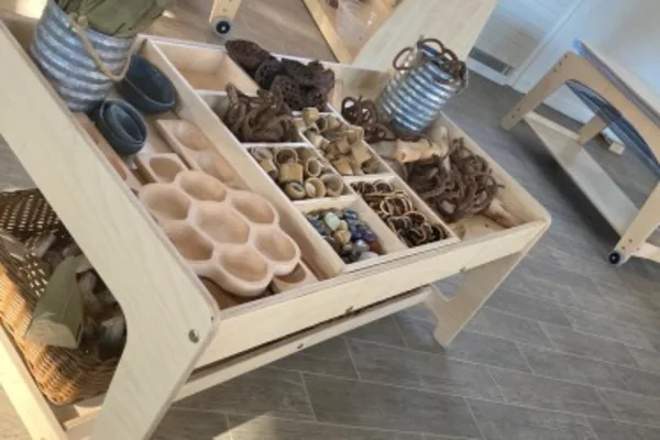 Low wooden tray organizer displays divided compartments of wooden scoops, beads, shells, coils and metal buckets, sitting on a tiled floor in a bright child-sized playroom with benches.