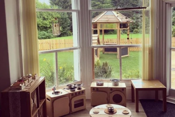 Wooden children's play kitchen and table are arranged by a bay window, set with toy dishes and utensils; beyond, a grassy yard contains a wooden gazebo and picket fence.