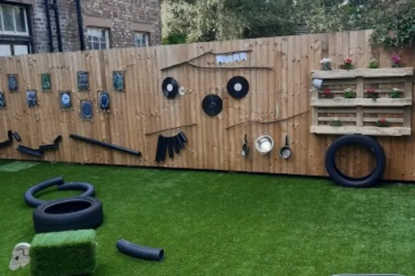 Wooden fence decorated with recycled items arranged like a smiling face (vinyl records, pots, pipes); artificial-turf yard in front with tires, turf blocks and a pallet planter.