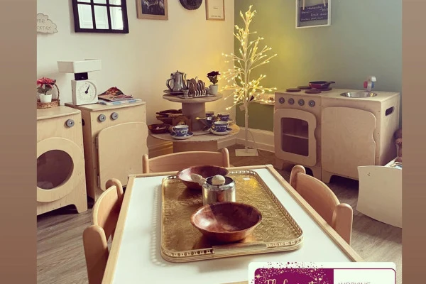 Wooden child-size table — set with a brass tray and wooden bowls — surrounded by wooden play-kitchen appliances and a small lit decorative tree in a pastel playroom. Text: small signs and a purple sticker are illegible.