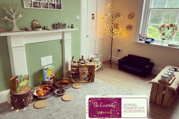 Child-sized play area of wooden crates, metal sensory bowls, and low furniture arranged on a rug, set against a pale green wall with a white mantel, lit tree, and window.