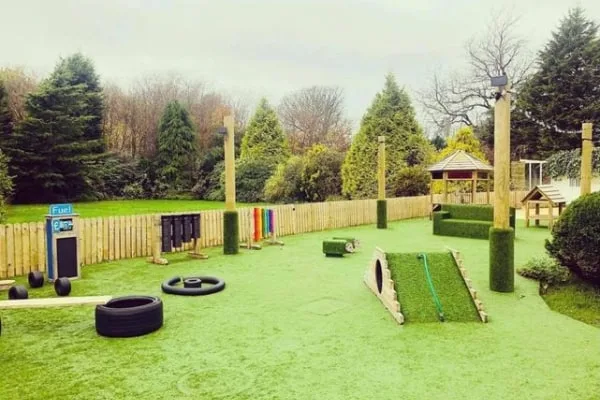 Artificial-turf playground ramp and tires form an obstacle course (object), arranged for children to climb and crawl (action), within a fenced yard surrounded by evergreen trees and a wooden gazebo (context). Text: "Fuel"