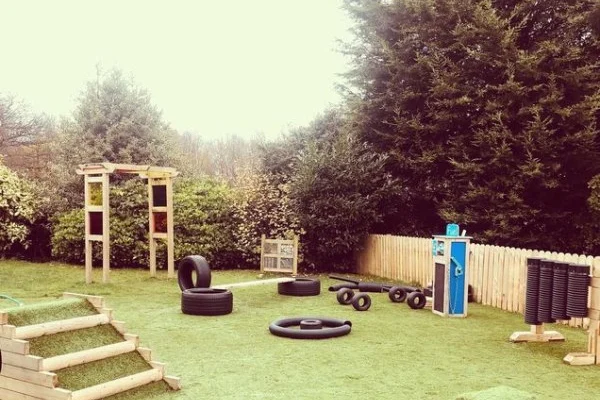 Playground equipment — stacked and scattered tires, balance beams, and a wooden climbing frame — sits arranged on artificial grass inside a fenced backyard bordered by tall evergreen trees.