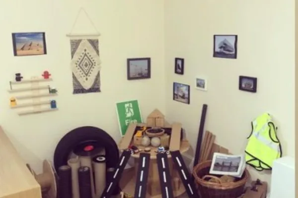 Cardboard toy road pieces converge on a small round table in a cluttered corner with framed photos, rolls of material and a high-visibility vest. Sign reads "Fire".