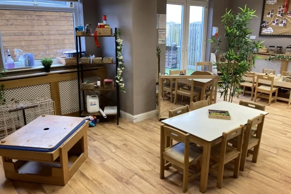 Small wooden table and chairs hold a box of crayons in a tidy preschool room with wooden floors, shelves, play stations, plants, and glass doors leading to an outdoor yard.